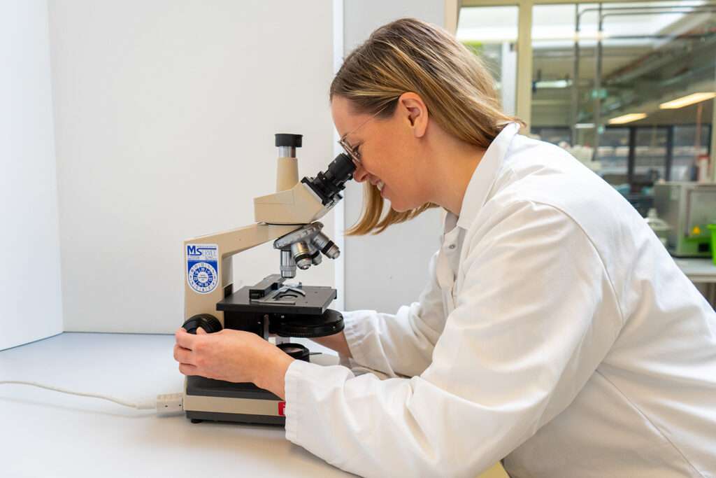 A person wearing a white lab coat is looking into a microscope in a bright laboratory, adjusting the focus knob, with lab equipment visible in the background—perfect for those interested in offene Stellen in scientific research.