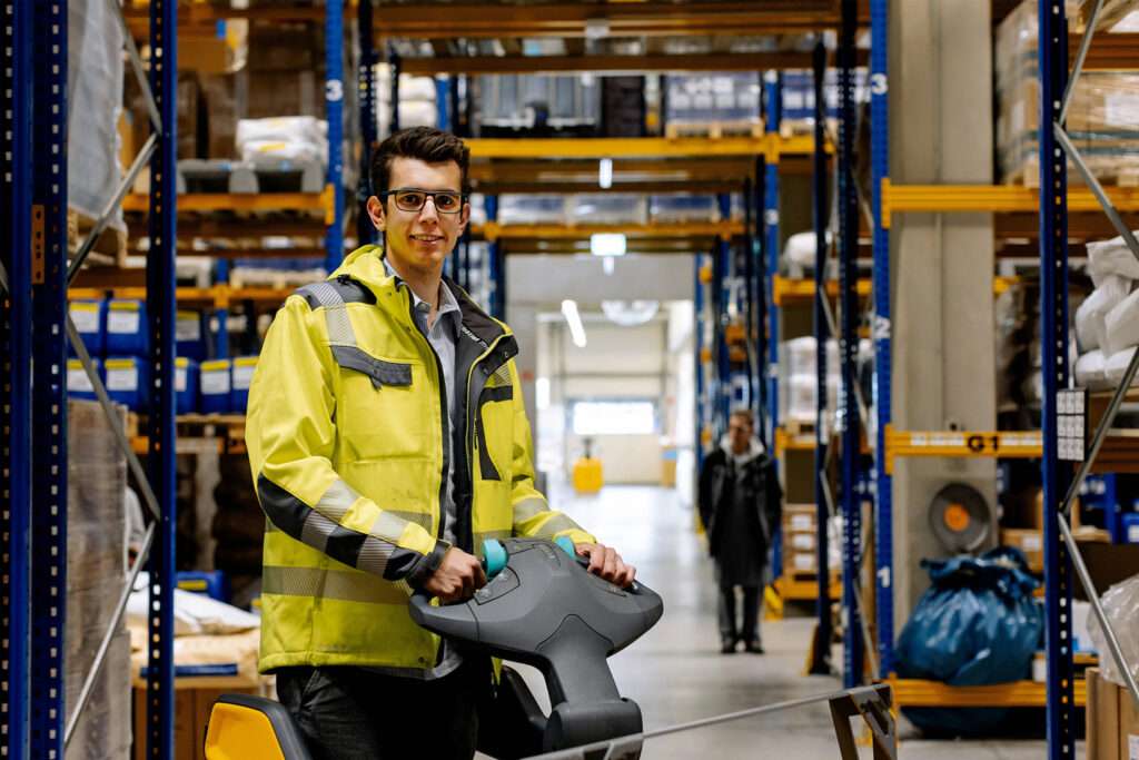 A man wearing safety glasses and a yellow high visibility jacket operates a pallet jack in a warehouse aisle, highlighting the active environment where offene Stellen for skilled workers may be available among shelves filled with boxes and supplies.
