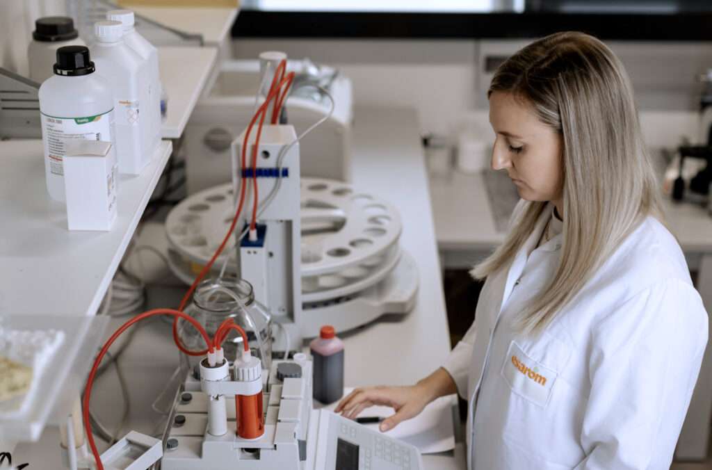 A woman in a white lab coat works with scientific equipment and chemicals on a laboratory bench, focusing on a machine with buttons and red tubes. Bottles and lab instruments are visible around her.