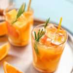 Two glasses filled with an orange coloured drink, ice cubes, orange slices, and garnished with rosemary sprigs. The vibrant display highlights the refreshing flavour, with extra orange wedges on a tray against a blue background.