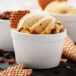 A scoop of ice cream with raisins in a white cup, featuring a unique flavour and garnished with pieces of waffle cone. Another cup and a waffle cone are visible in the background on a dark surface.