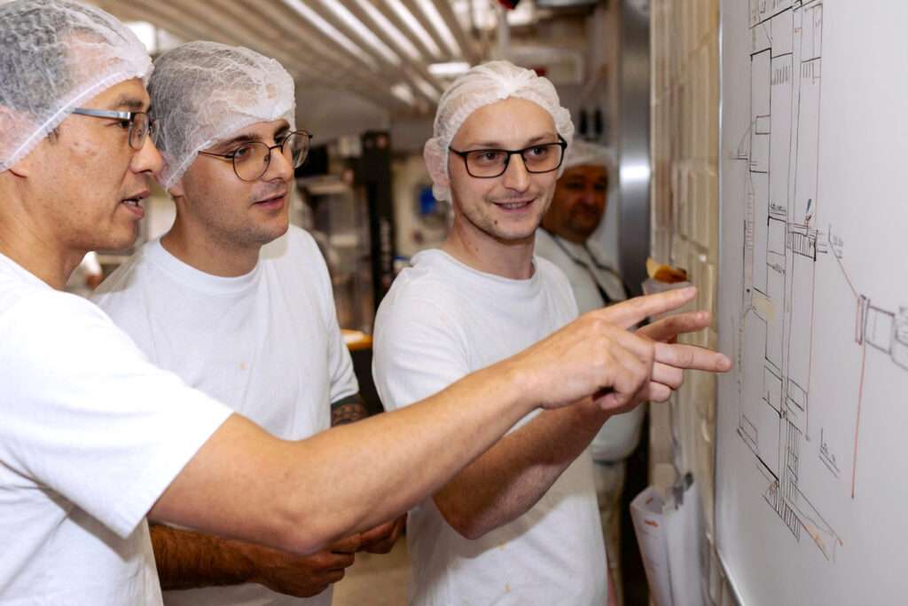 Three people wearing white T shirts and hairnets are standing together, looking at and discussing a technical drawing or diagram on a whiteboard in a brightly lit room, possibly reviewing offene Stellen for improvement.