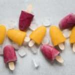 Eight popsicles, four yellow and four purple, are scattered on a light gray surface among several ice cubes. Each wooden stick holds a treat starting to melt, hinting at their refreshing flavour.