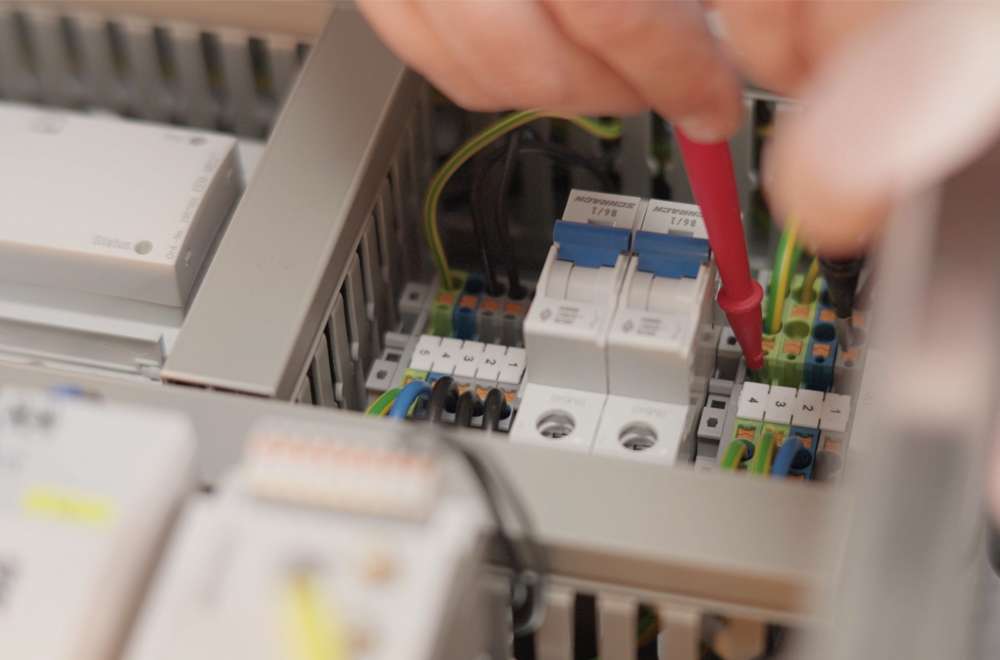 A close up of a person using a red screwdriver to adjust wiring inside an electrical control panel, showing various wires and circuit breakers.