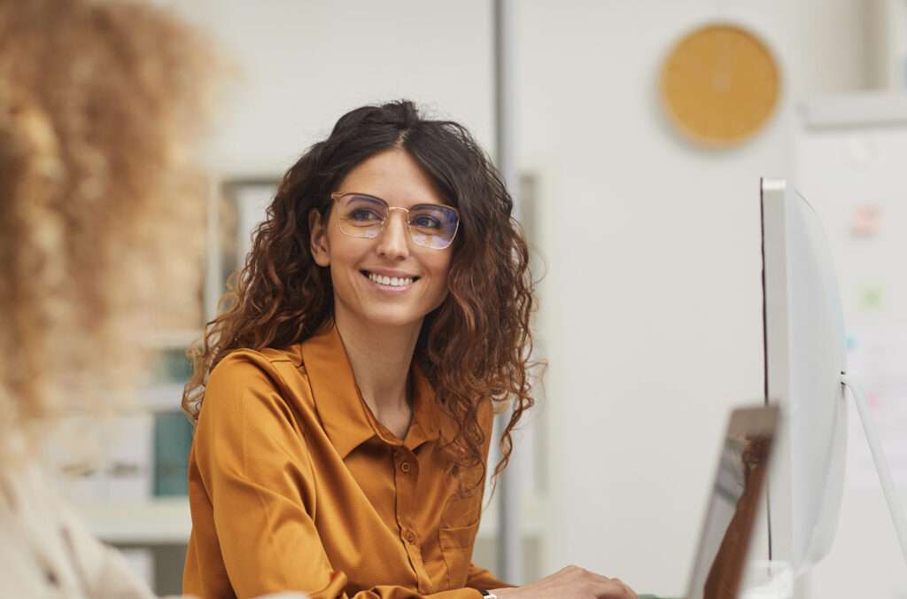 A woman with curly brown hair and glasses, wearing a mustard yellow blouse, smiles while sitting at a desk with a computer monitor. Another person is partially visible in the foreground.