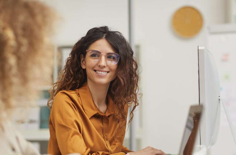 A woman with curly hair and glasses, wearing a mustard-colored blouse, smiles while sitting at a desk with a computer monitor. Another person is slightly visible in the foreground. The setting appears to be an office.