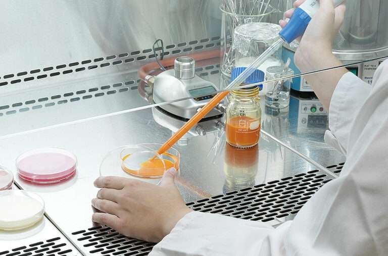 A scientist in a lab coat uses a pipette to transfer orange liquid into a petri dish under a fume hood, with laboratory equipment and glassware visible in the background.