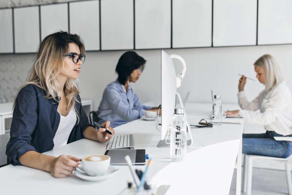 Three women working at desks in a modern, bright office space. One woman in the foreground holds a cup of coffee and a pen, with a computer monitor in front of her. The other two women work at their computers in the background.