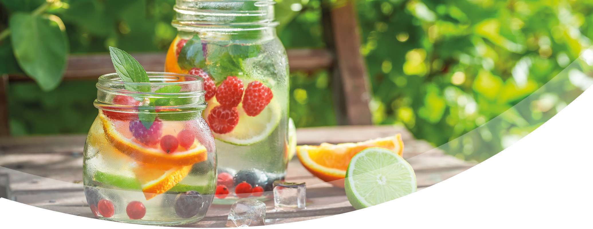 Zwei mit Wasser, Orangenscheiben, Limetten, Beeren und Minzblättern gefüllte Gläser stehen auf einem Holztisch im Freien, umgeben von Grünzeug und Sonnenlicht.