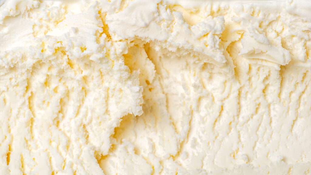 Close up of creamy vanilla ice cream with a smooth, textured surface and visible scoop marks, highlighting its rich flavour. The ice cream is pale yellow and white in color.