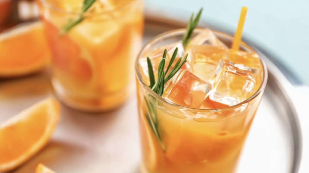 A close up of a glass filled with iced orange drink, bursting with citrus flavour, garnished with orange slices and a sprig of rosemary, with another similar glass and fresh orange wedges in the background on a tray.