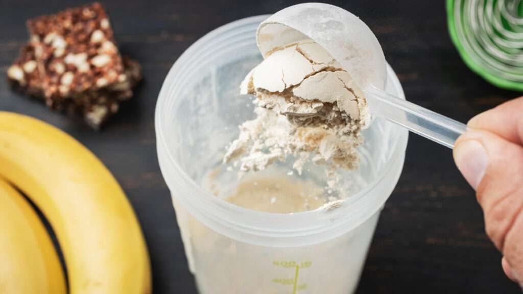 A hand holds a scoop of flavoured protein powder over a shaker cup, with a banana and a snack bar next to it on a dark surface.