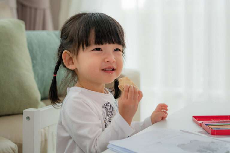 A young child with pigtails smiles while eating a wafer biscuit featured in Confectionery News. She sits at a white table with colored pencils and paper in front of her, and a sofa with cushions is in the background.