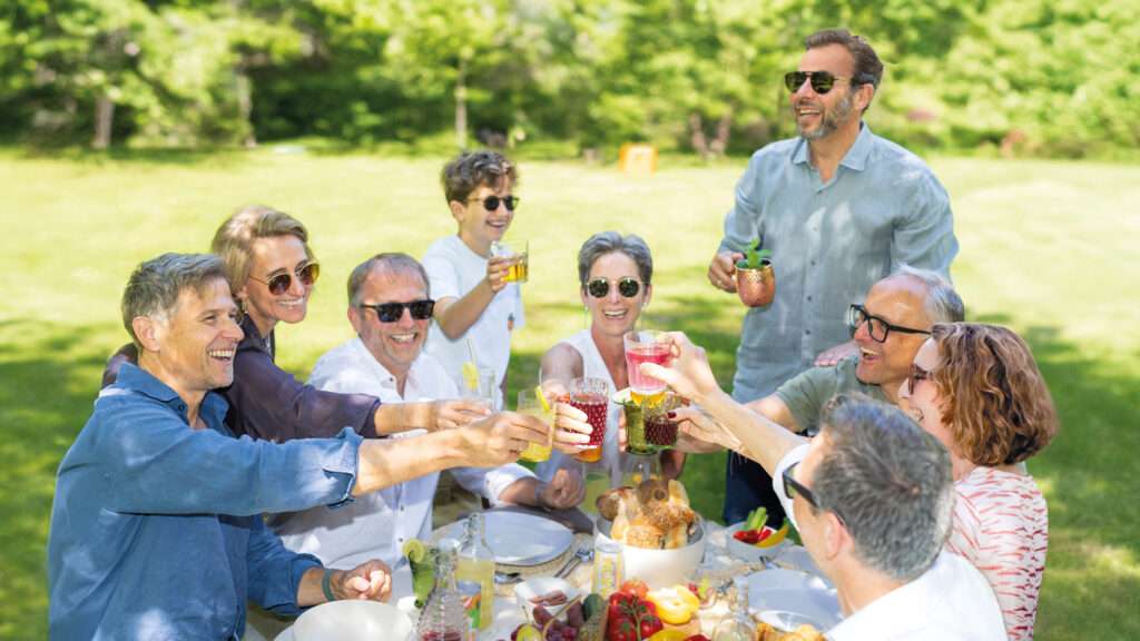 A group of adults sitting outdoors at a picnic table, raising glasses and smiling, surrounded by food and greenery on a sunny day—capturing the warmth and camaraderie reminiscent of gatherings inspired by drinktec 2025.