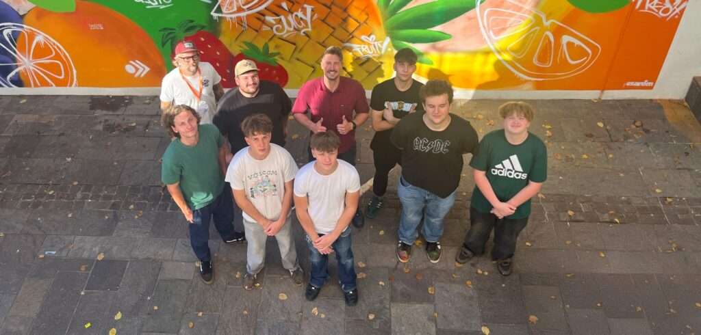 Eine Gruppe von zehn Personen steht lächelnd auf einem Steinweg vor einem farbenfrohen Wandgemälde mit Obstmotiven beim esarom Lehrlingsevent. Das Sonnenlicht wirft Schatten auf den Boden, während alle, leger gekleidet, in die Kamera blicken.
