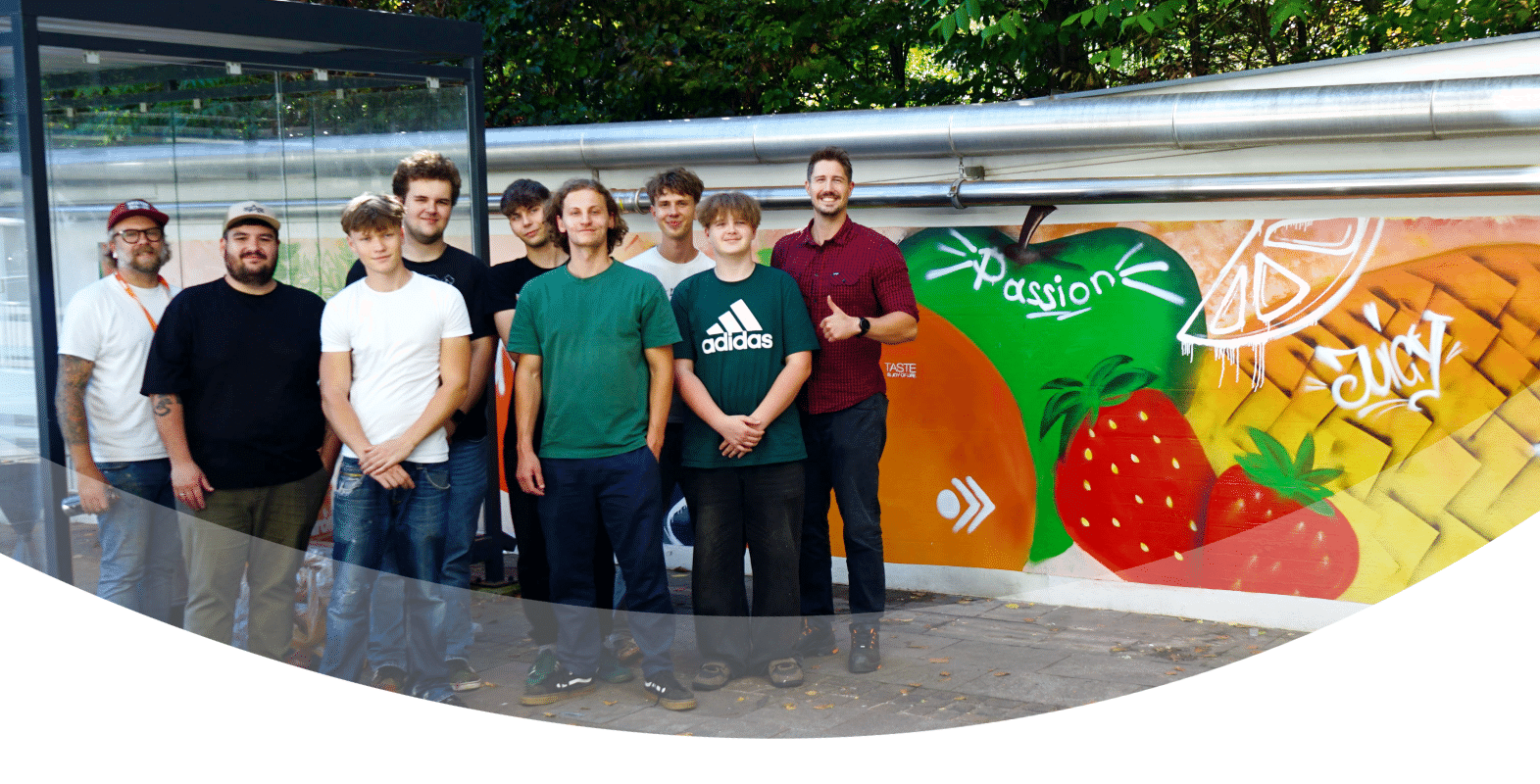 Eine Gruppe von zehn Personen steht lächelnd vor einem farbenfrohen Wandbild, auf dem Früchte sowie die Wörter „Passion“ und „Juicy“ zu sehen sind. Im Hintergrund sind Bäume und eine Glaskonstruktion zu erkennen, die einen lebhaften Moment einer esarom Lehrlingsveranstaltung einfangen.