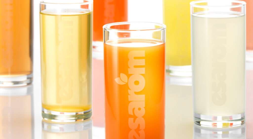 Six glasses of colourful fruit juices in shades of orange, yellow, and clear are arranged on a reflective surface, each capturing the vibrant essence of fruit flavour. The word “esarom” is printed in white on every glass.