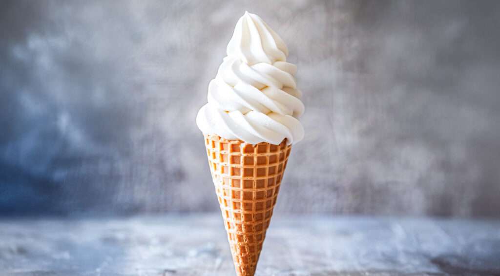 A close up of a waffle cone filled with a tall swirl of vanilla soft serve ice cream, showcasing its classic flavour against a blurred gray background.