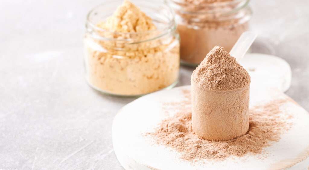 A scoop of brown protein powder, known for its rich flavour, sits on a white surface with jars of yellow and brown powders blurred in the background.