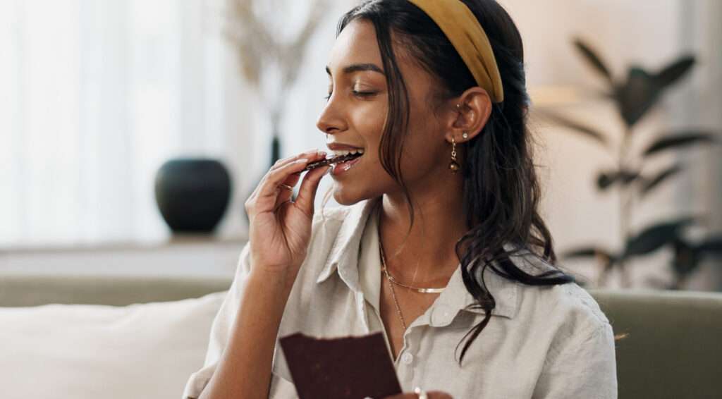 A woman with a yellow headband smiles as she enjoys the rich flavour of a piece of chocolate while sitting indoors, with blurred plants and soft light in the background.