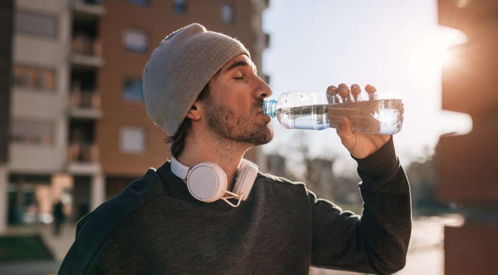 A man wearing a gray beanie and headphones around his neck drinks water from a plastic bottle while standing outdoors, enjoying the refreshing flavour as sunlight shines in the background.
