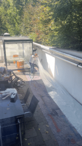 Eine Person in einem weißen Hemd und einer roten Mütze breitet eine große Plastikplane auf einer Terrasse neben einer weißen Wand aus; in der Nähe stehen Tische, Stühle und orangefarbene Fässer – offensichtlich werden gerade die Vorbereitungen für die „esarom“ Lehrlingsveranstaltung getroffen. Im Hintergrund sind Bäume und Sonnenlicht zu sehen, was eine einladende Kulisse bildet, die wahrscheinlich das Interesse der Medien wecken wird.