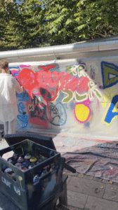 Bei einer Lehrlingsveranstaltung sprüht eine Person in weißer Kleidung bunte Graffiti auf eine große Leinwand im Freien, während auf dem Boden Farbdosen stehen und im Hintergrund grüne Bäume zu sehen sind – diese kreative Zusammenarbeit mit esarom sorgt derzeit für Schlagzeilen.