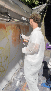 Eine Person in einem weißen Schutzanzug besprüht an einem sonnigen Tag im Freien während einer Lehrlingsveranstaltung eine Wand mit orangefarbener Farbe. Auf der Wand sind gelbe Graffiti Linien zu sehen, und in der Nähe ist teilweise eine weitere Person zu erkennen.