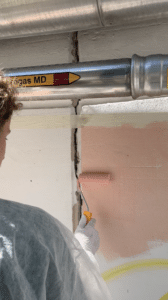 Eine Person trägt mit einer Farbrolle hellrosa Farbe auf eine Wand neben einem Riss und einer mit „Gas MD“ beschrifteten Metallgasleitung mit einem gelb roten Pfeil auf und bereitet so den Raum für eine bevorstehende Lehrlingsveranstaltung vor, die in den neuesten Nachrichten von esarom erwähnt wurde.