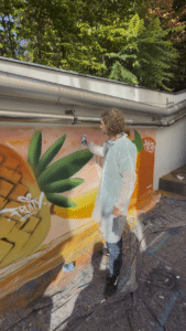 Im Rahmen einer Lehrlingsveranstaltung von esarom sprüht eine Person in einem weißen Schutzanzug ein farbenfrohes Wandbild einer Ananas mit der Aufschrift „FRUIT“ an eine Außenwand, über der grüne Bäume zu sehen sind – ein Bild, das die in den jüngsten Nachrichten beschriebene lebhafte Stimmung einfängt.