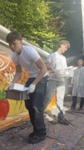 Zwei junge Männer tragen Behälter vor einem farbenfrohen Wandbild mit einer Tomate beim Lehrlingsevent von esarom. Der eine trägt Handschuhe und ein weißes Hemd, der andere ein gemustertes Hemd. Im Hintergrund stehen zwei weitere Personen, von denen eine einen Laborkittel trägt. Sonnenlicht fällt durch die Bäume über ihnen und schafft eine stimmungsvolle Kulisse für dieses spannende News Update.