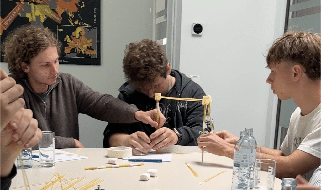 Drei junge Leute sitzen an einem Tisch und bauen aus Spaghetti und Marshmallows einen Turm – dabei arbeiten sie bei einer lebhaften Lehrlingsveranstaltung bei esarom eng zusammen. Wasserflaschen, Marshmallows und Spaghetti Stangen liegen verstreut auf dem Tisch, während sie gemeinsam an dieser kreativen Herausforderung arbeiten.